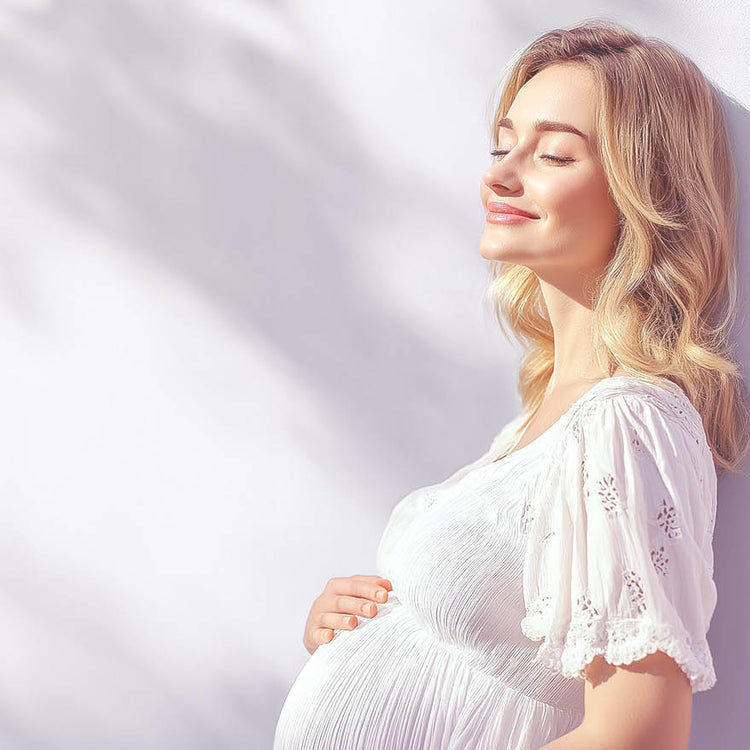 Eine Junge schwangere Frau lehnt sich an eine helle Wand und wird von der Sonne angestrahlt.